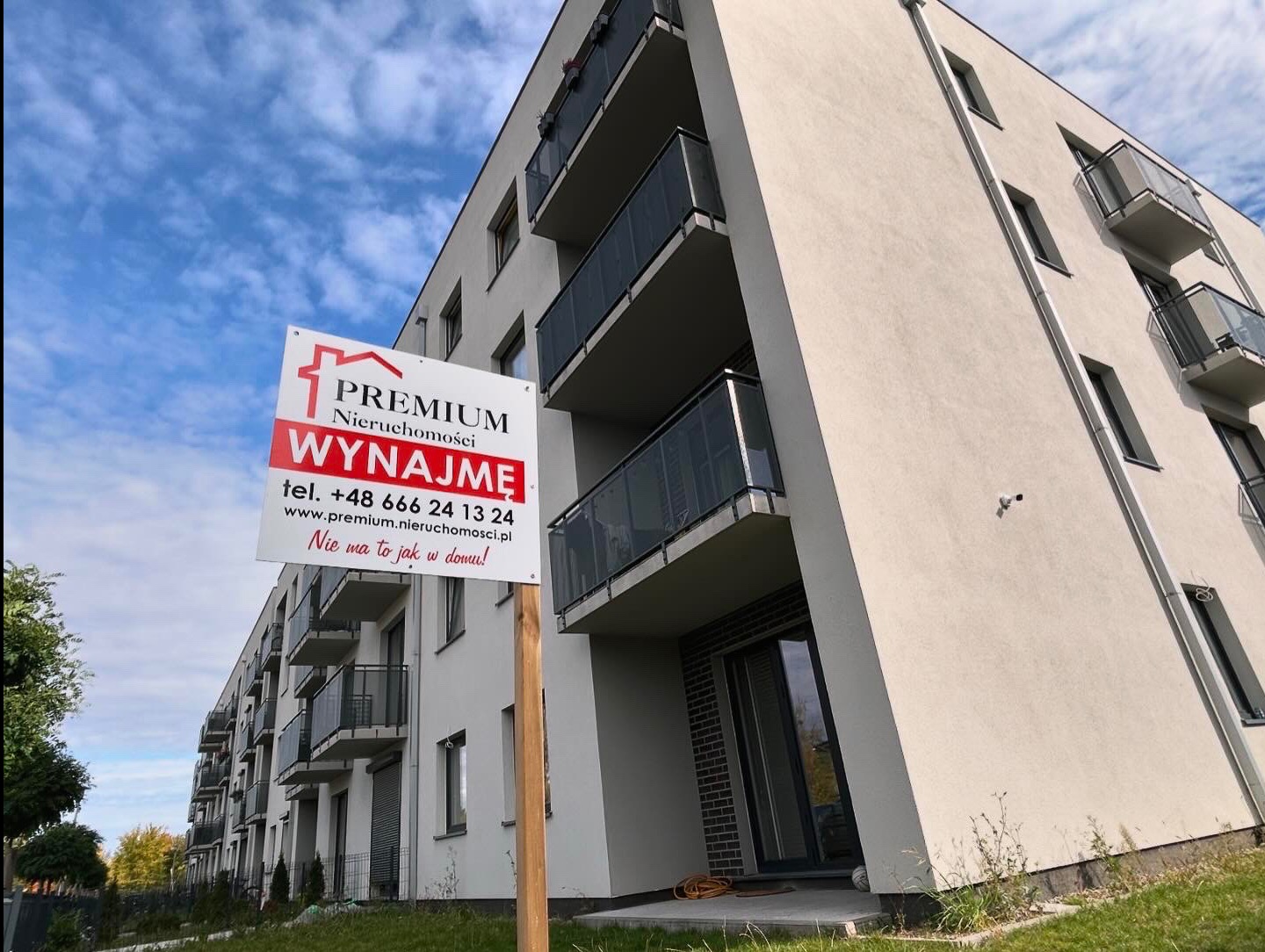 Nowoczesny budynek wielorodzinny z balkonami, przed którym umieszczono tablicę reklamową agencji nieruchomości z napisem 'Wynajmę' na tle błękitnego nieba.