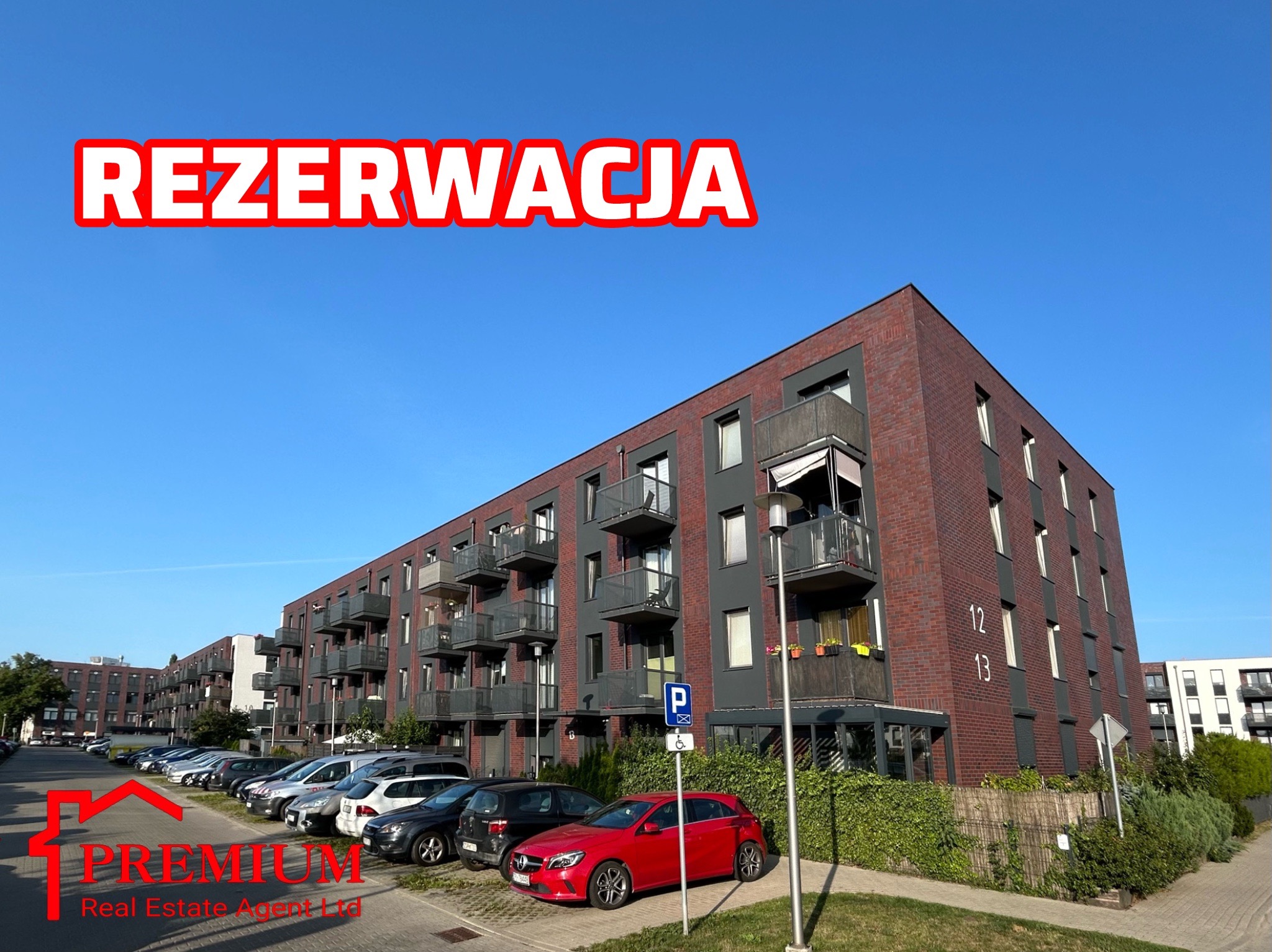 Nowoczesny apartamentowiec z cegły z balkonami i parkingiem dla mieszkańców, oznaczony napisem 'Rezerwacja' na niebie.