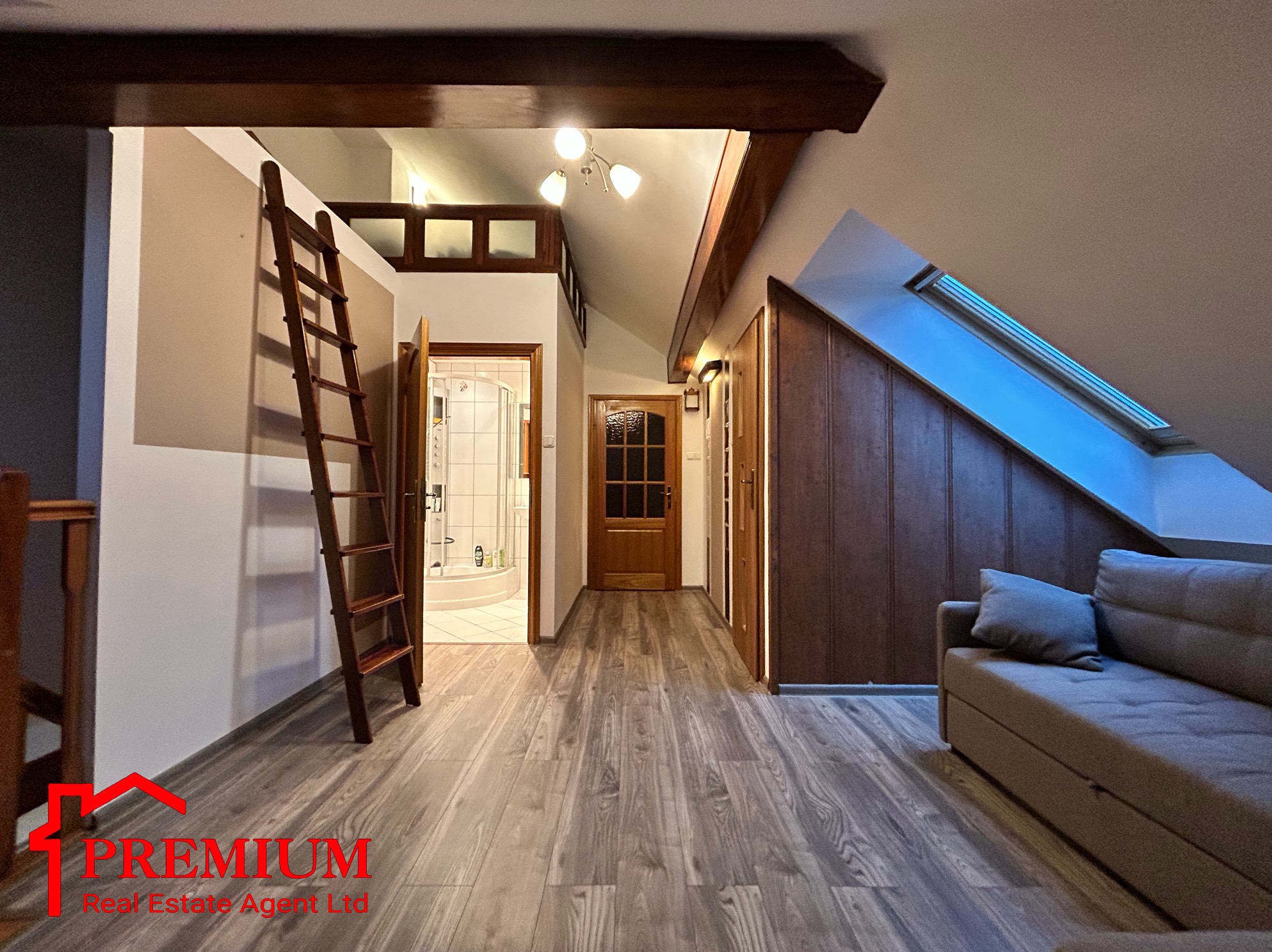 Poddasze z drewnianymi belkami, schodami drabinkowymi, łazienką z prysznicem w tle, drewnianą podłogą i szarą sofą, oświetlone światłem dziennym z okna dachowego.