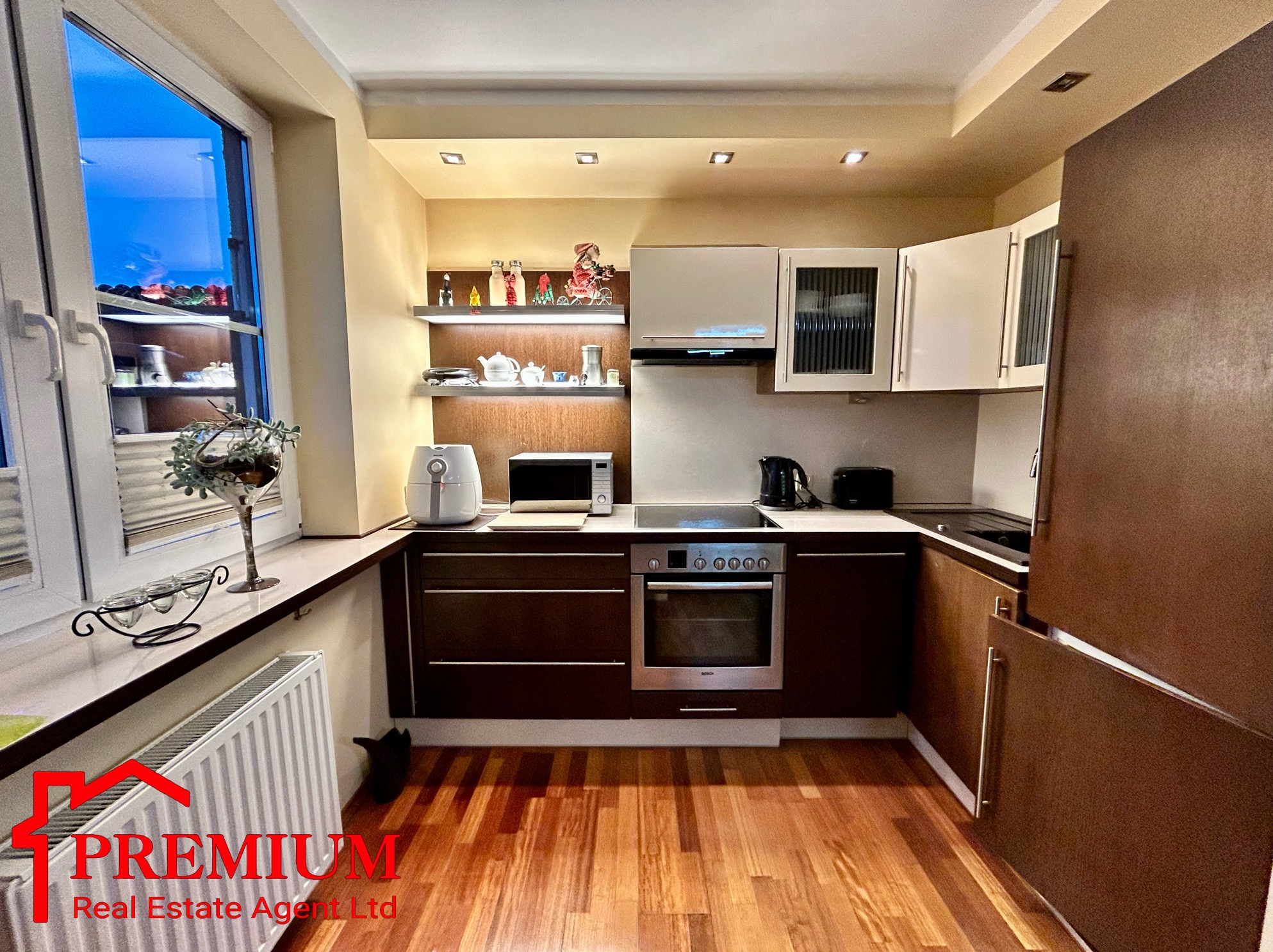 Nowoczesna kuchnia z drewnianą podłogą, brązowymi szafkami dolnymi i białymi górnymi, z widocznym oknem i dekoracjami na parapecie oraz logo agencji nieruchomości w lewym dolnym rogu.