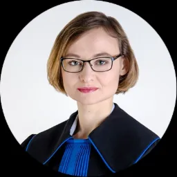 Portret kobiety w todze prawniczej z niebieskimi plisami, w okularach, na jasnym tle.