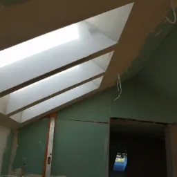 Poddasze w Olsztynie z oknami dachowymi i ścianami pokrytymi płytami gipsowo-kartonowymi przed szpachlowaniem, widoczny poziomica i przewody elektryczne.