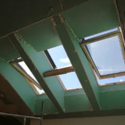 Montaż okien dachowych w poddaszu z widocznymi płytami gipsowo-kartonowymi i pianką montażową, widok na błękitne niebo