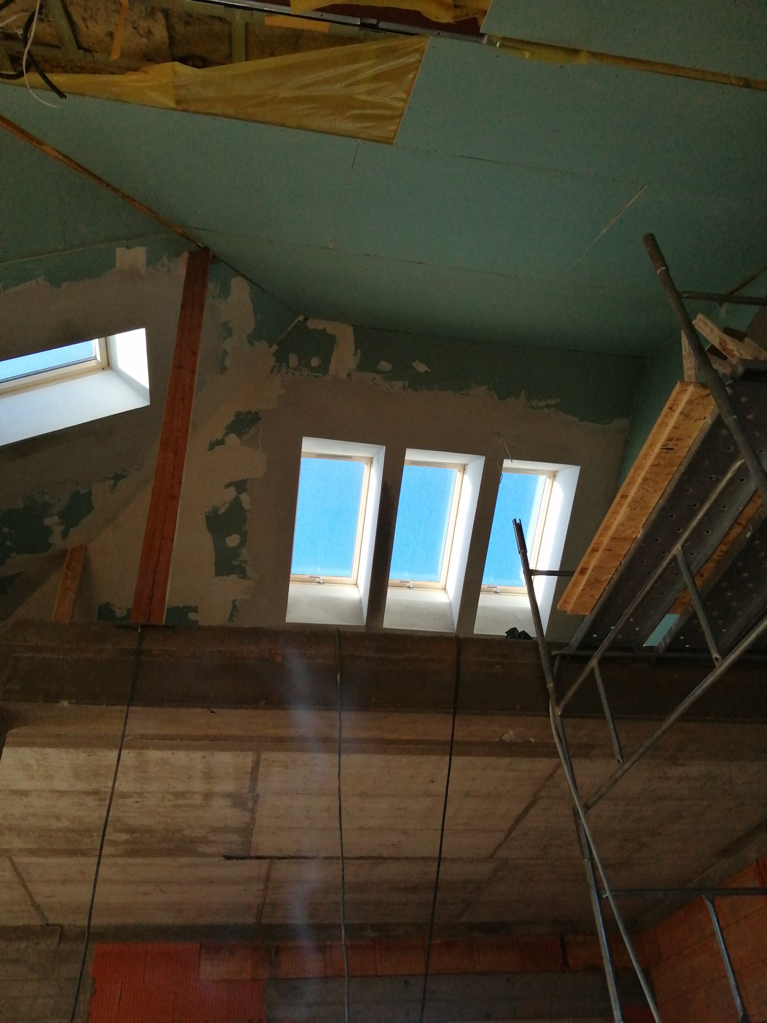 Wnętrze poddasza w trakcie prac wykończeniowych: trzy okna dachowe w ścianie szczytowej, widoczne rusztowanie, surowy betonowy strop, częściowo położona płyta gipsowo-kartonowa na suficie.