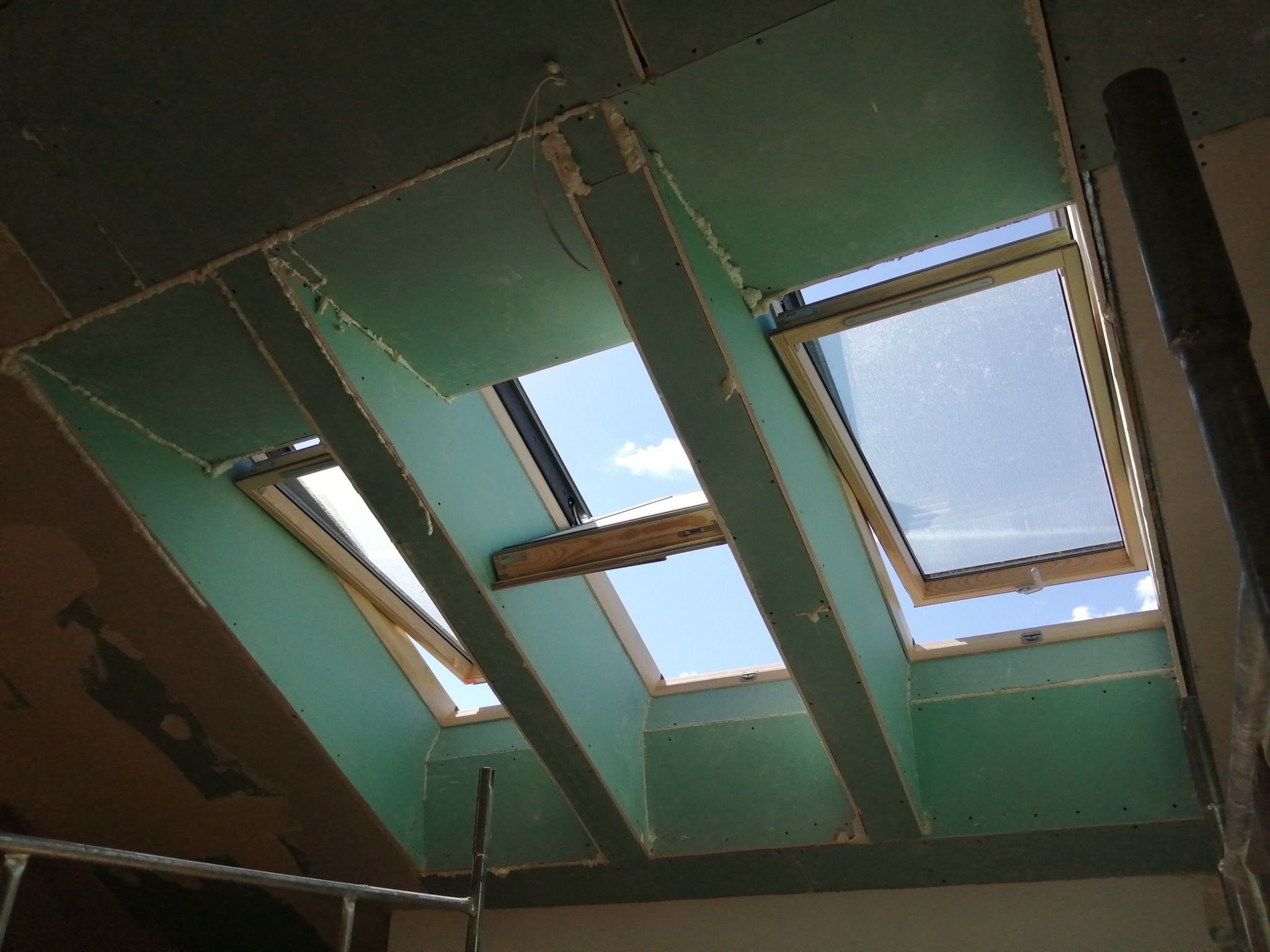 Montaż okien dachowych w poddaszu z widocznymi płytami gipsowo-kartonowymi i pianką montażową, widok na błękitne niebo