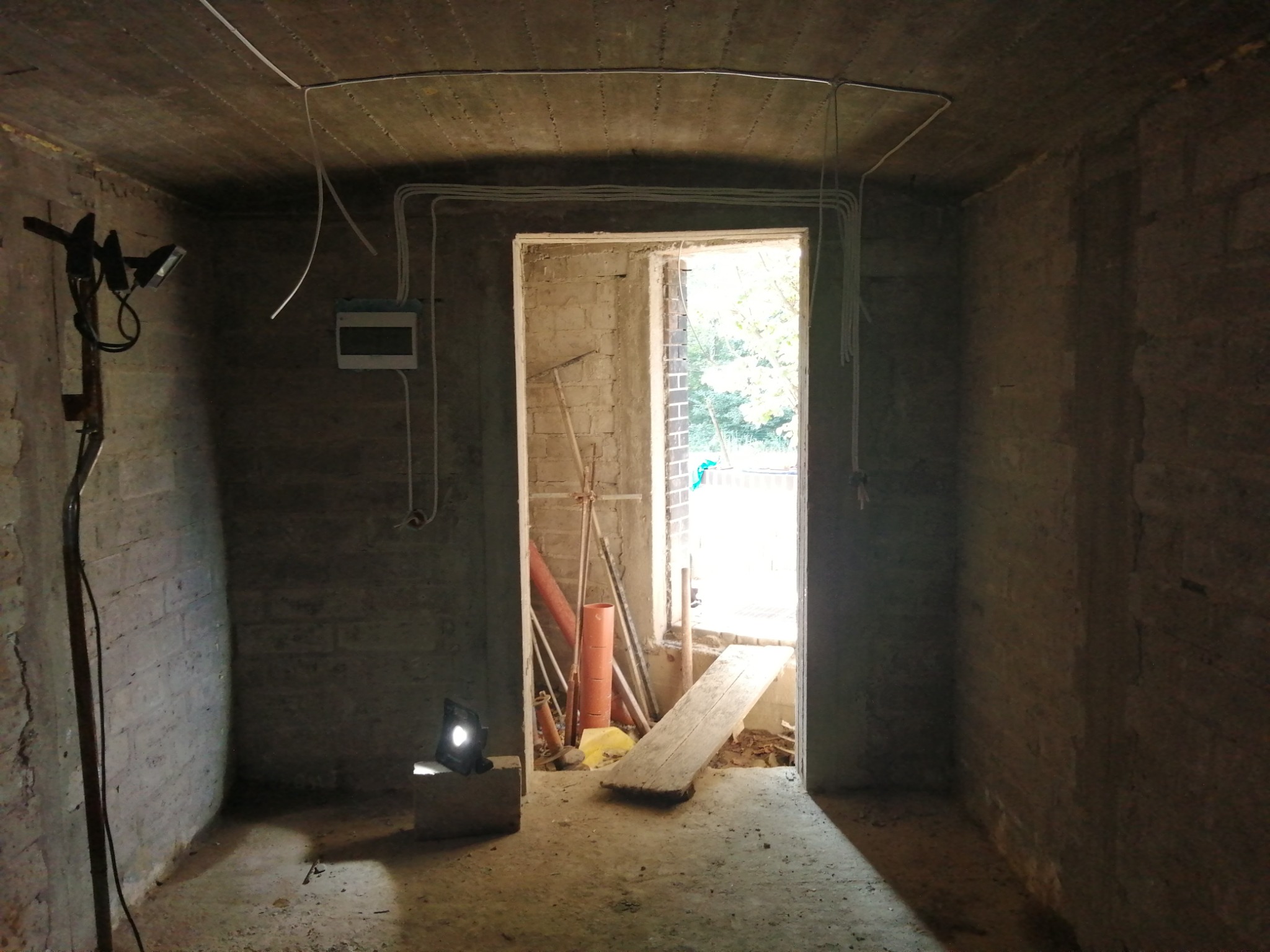 Surowe wnętrze w trakcie prac przygotowawczych: widoczne przewody elektryczne na suficie, ściany z surowej cegły, prowizoryczne oświetlenie halogenowe i narzędzia w przejściu drzwiowym.