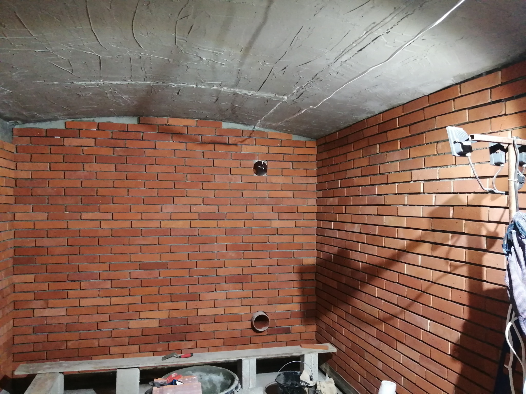 Wnętrze w trakcie wykończenia z ceglanymi ścianami, surowym betonowym sufitem i widocznymi instalacjami, oświetlone lampą budowlaną.