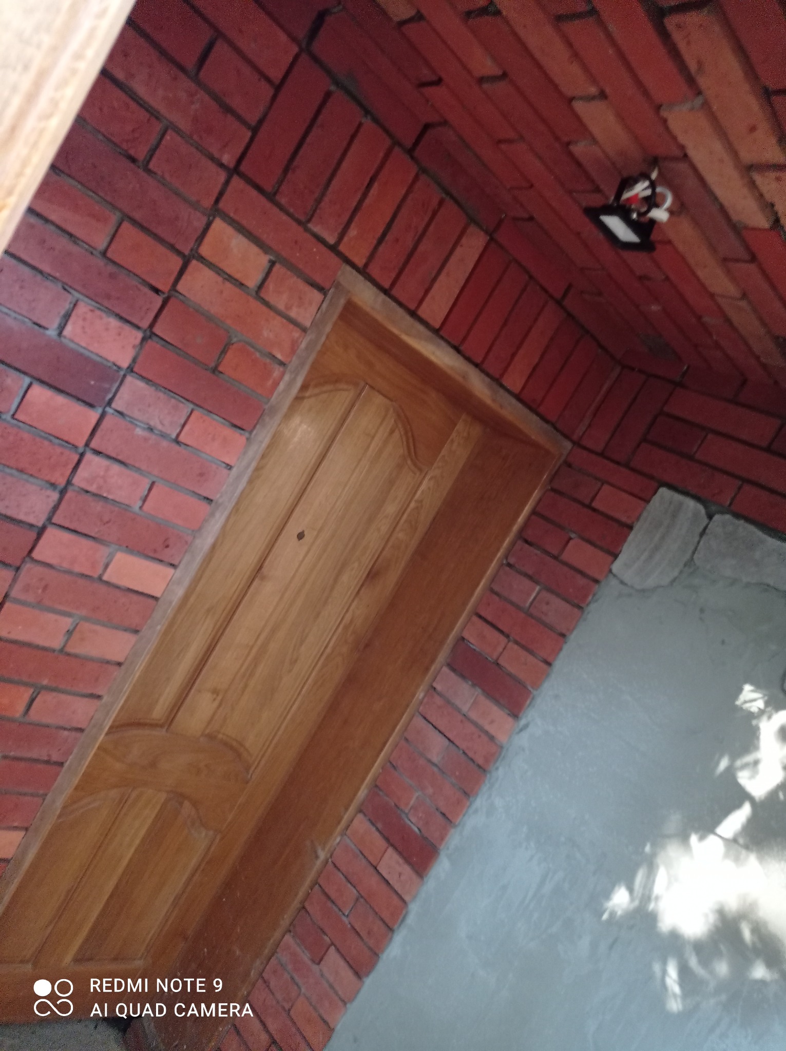 Drewniane drzwi w czerwonej ceglanej wnęce, widoczne światło sufitowe i szara podłoga z odbiciem światła.