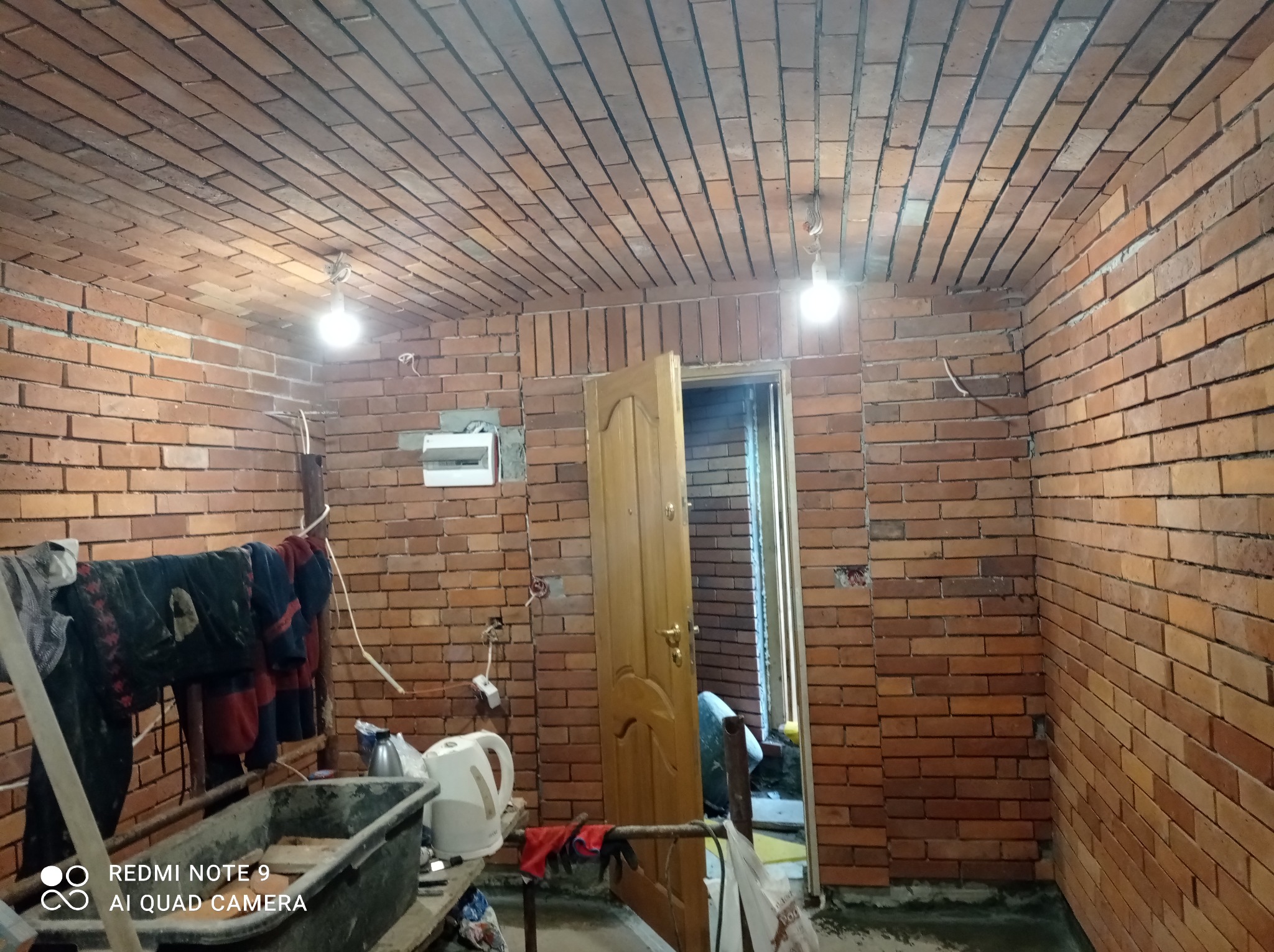 Ceglana ściana i sufit w pomieszczeniu w trakcie remontu, z widocznymi niedokończonymi fragmentami, wiszącymi kablami, otwartymi drzwiami i prowizorycznym wieszakiem z ubraniami.