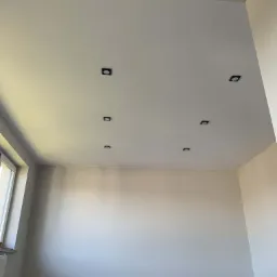 Wnętrze pokoju z zamontowanymi, czarnymi, kwadratowymi lampami sufitowymi typu spot w rzędach, widoczne gniazdka elektryczne na ścianie i zwisające przewody na podłodze.