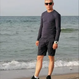 Młody mężczyzna w okularach przeciwsłonecznych, ciemnej koszulce z długim rękawem i szortach, stojący na piaszczystej plaży z falami morskimi w tle.