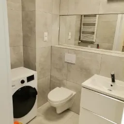 Nowoczesna łazienka z szarymi płytkami imitującymi beton, podwieszana toaleta, biała szafka z umywalką, pralka i żółty kosz z mopami w rogu.