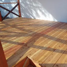 Świeżo wybudowany drewniany taras z balustradą, deski ułożone równolegle, widoczne słoje drewna i cień rzucany przez balustradę na białą ścianę w tle.