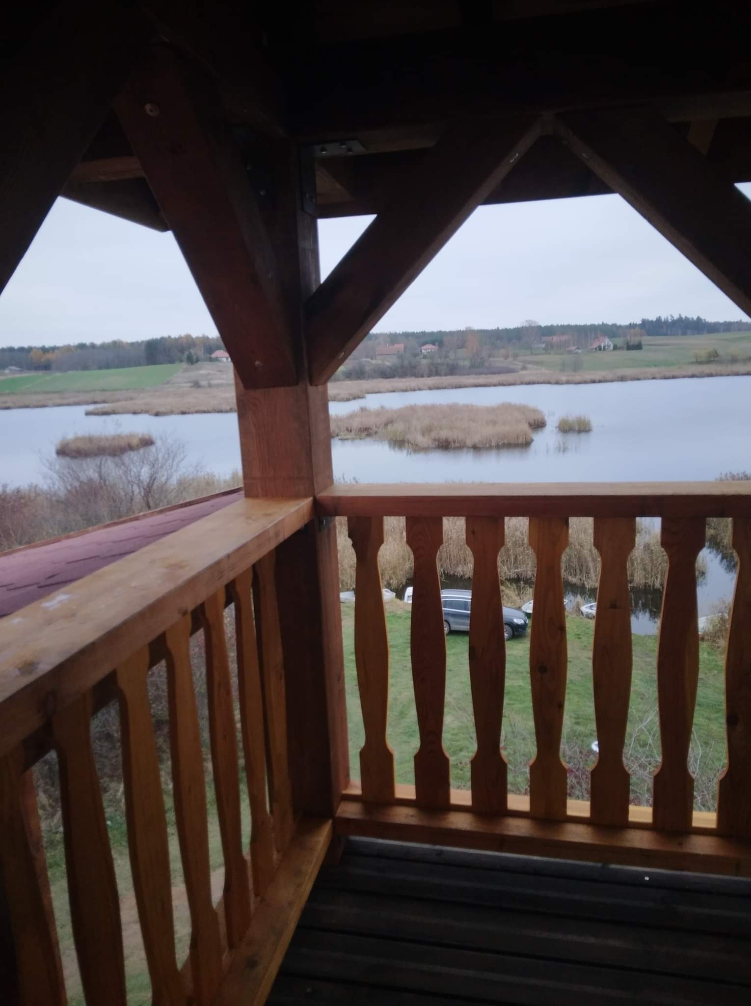 Drewniana balustrada z widokiem na jezioro z trzcinowymi wyspami i brzegiem, konstrukcja z belek, widoczny samochód i łódka na brzegu w tle.