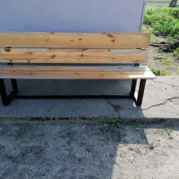 Drewniana ławka z metalowymi nogami, ustawiona na betonowym podłożu przed szarą ścianą budynku, z widocznymi śladami użytkowania i otoczeniem z roślinnością.