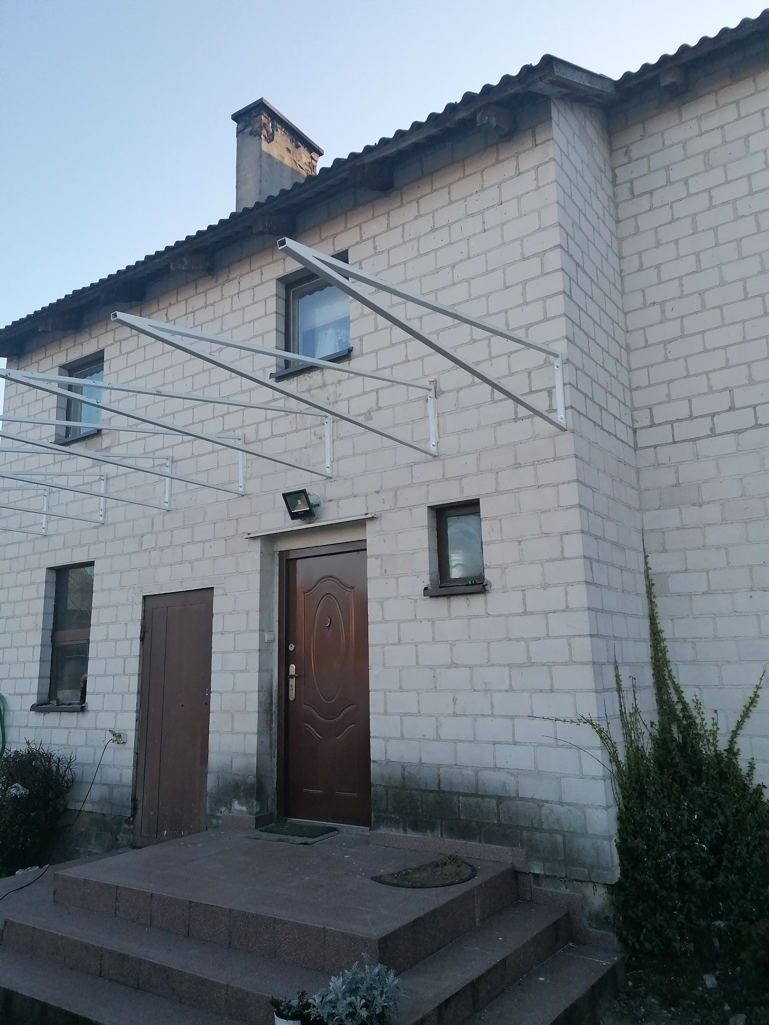 Biały budynek z cegły z zainstalowaną metalową konstrukcją nad wejściem, prawdopodobnie przygotowaną pod zadaszenie.