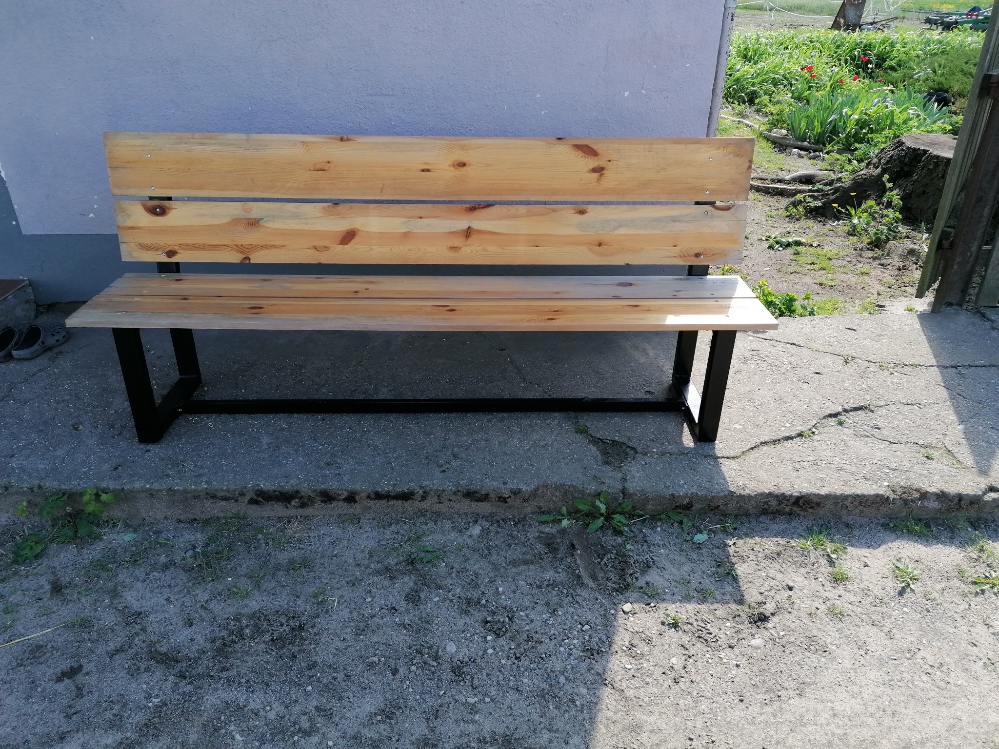 Drewniana ławka z metalowymi nogami, ustawiona na betonowym podłożu przed szarą ścianą budynku, z widocznymi śladami użytkowania i otoczeniem z roślinnością.