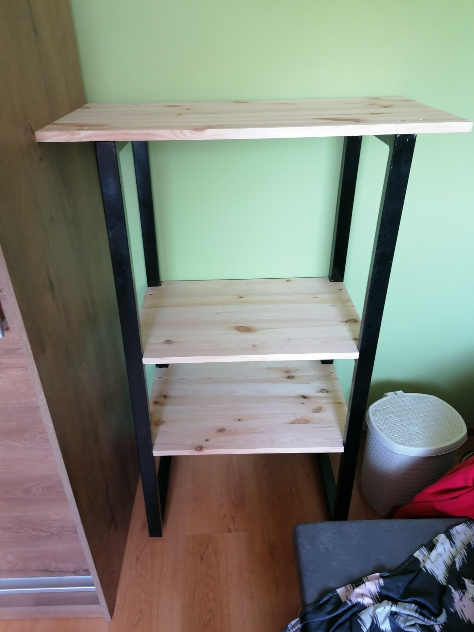 Trzypoziomowy regał z jasnego drewna i czarnych metalowych nóg, stojący przy zielonej ścianie, widoczny fragment szafy i kosza na pranie.