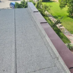 Widok z góry na pokrycie dachu papą z widocznymi łączeniami i brązowym obrzeżem, z zielonym trawnikiem i drzewami w tle.