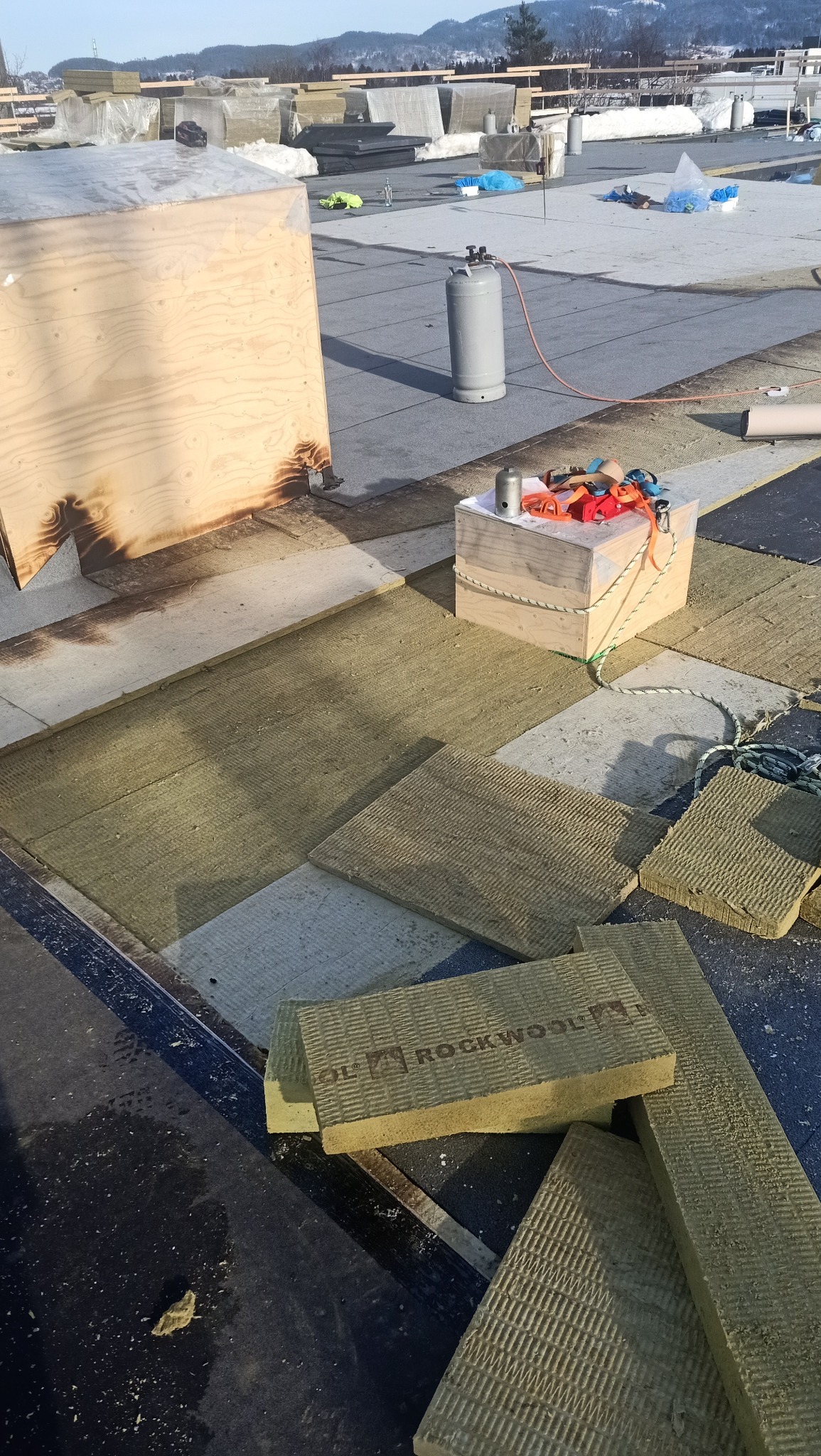 Montaż izolacji Rockwool na dachu płaskim, widoczny palnik gazowy, skrzynka narzędziowa i uszkodzona skrzynia z wypaleniami.