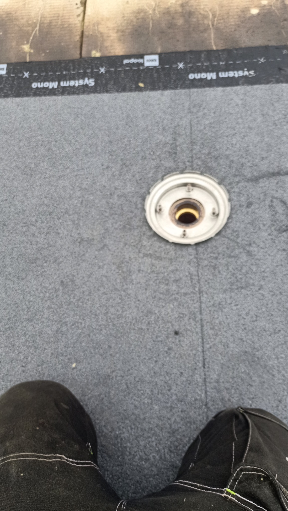 Szary materiał hydroizolacyjny z wbudowanym odpływem dachowym, widoczne spodnie robocze osoby wykonującej prace.