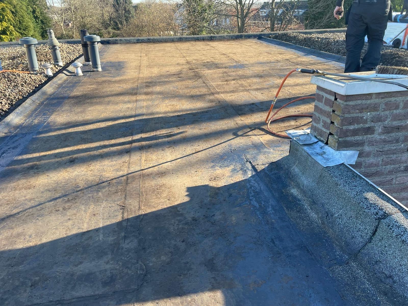 Płaski dach pokryty papą termozgrzewalną przed obróbkami blacharskimi komina, widoczne kominki wentylacyjne i narzędzia dekarskie