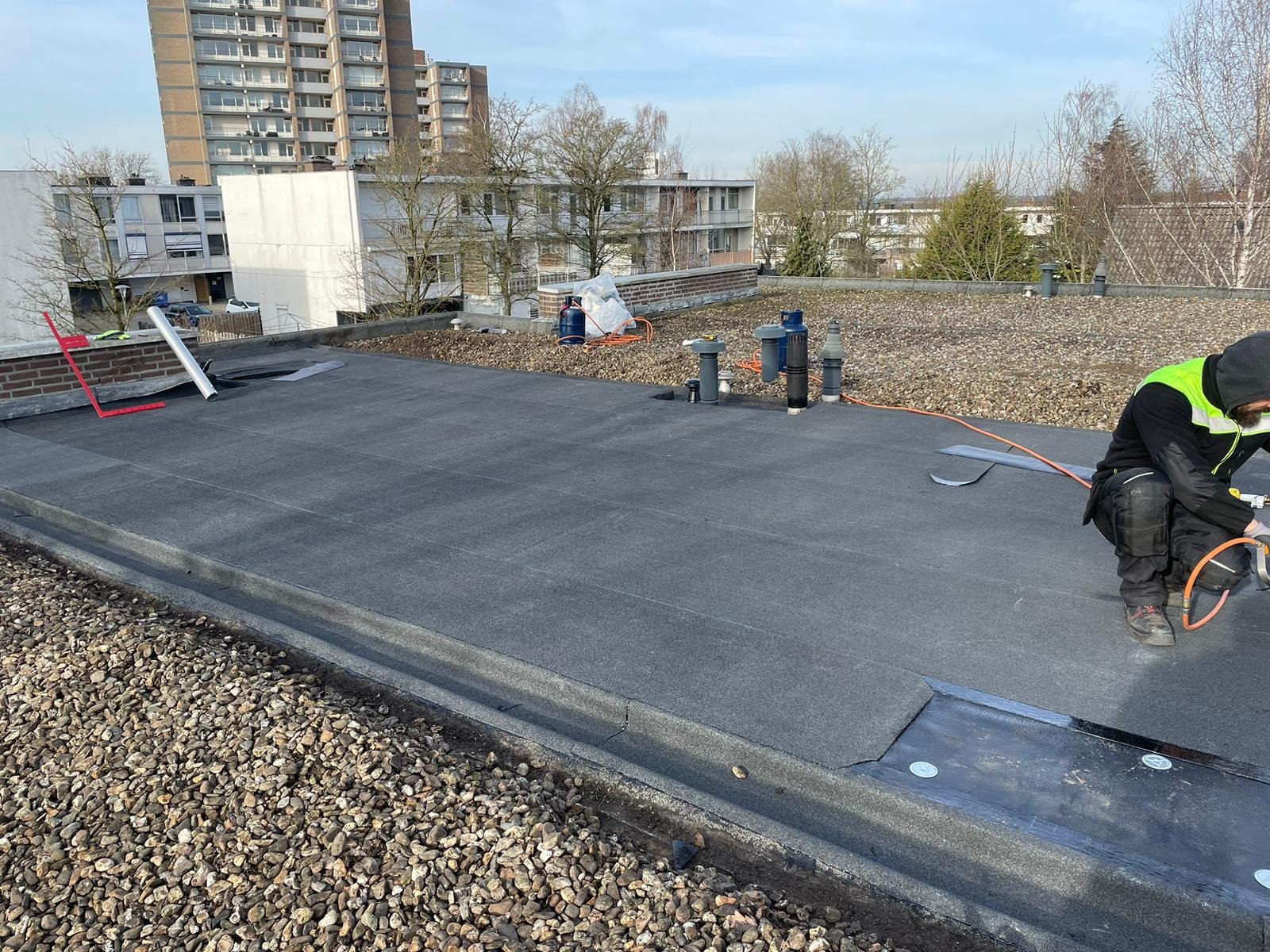 Pracownik w odzieży roboczej uszczelnia połączenie papy termozgrzewalnej na płaskim dachu budynku mieszkalnego, widoczne narzędzia, butle gazowe i elementy instalacji.