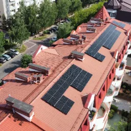 Widok z góry na budynek mieszkalny z czerwonym dachem, na którym zamontowano panele słoneczne w kilku sekcjach, otoczony zielenią i zabudową miejską.