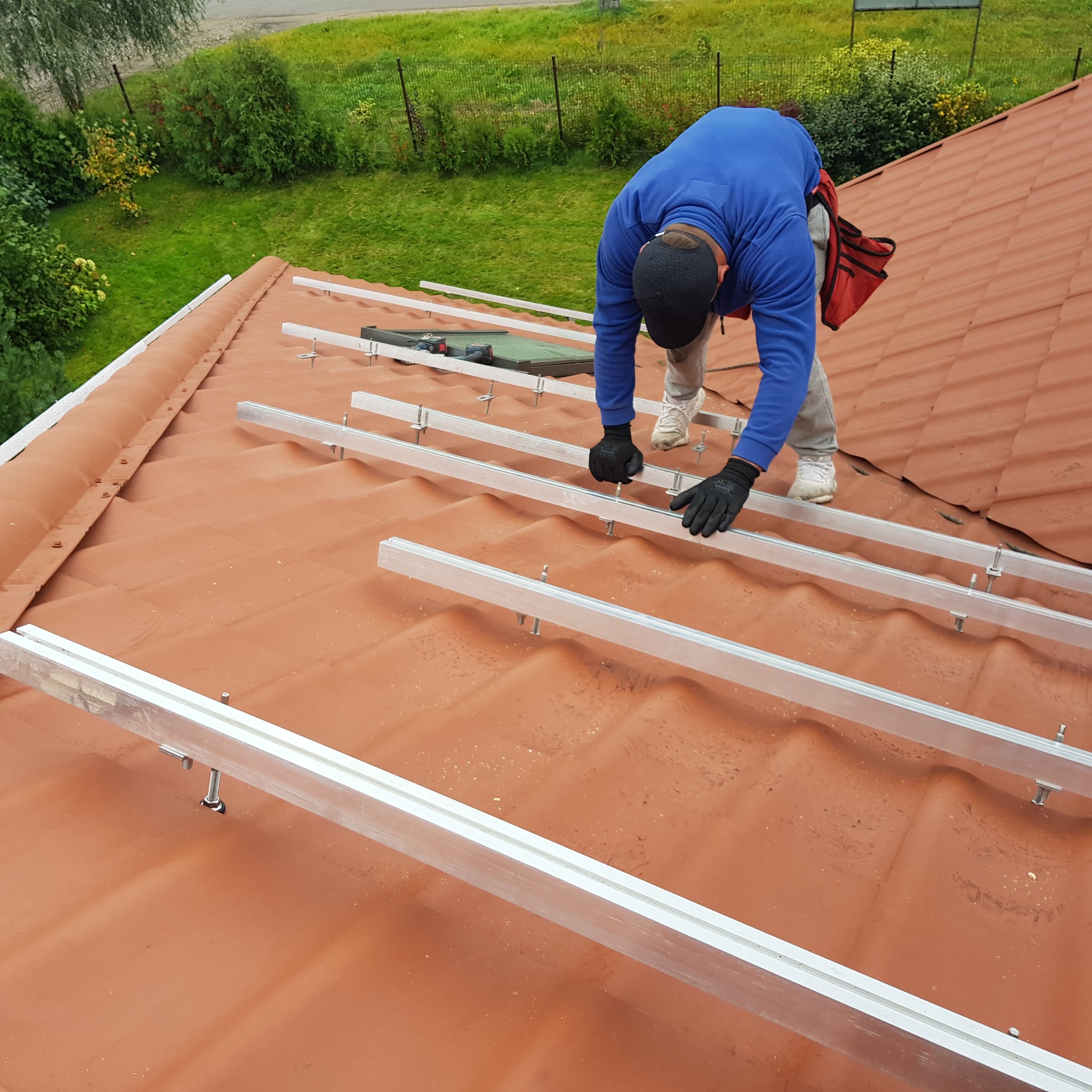 Montaż konstrukcji wsporczej z aluminiowych profili na dachu z dachówki ceramicznej pod panele słoneczne, widoczny monter w niebieskiej bluzie i czapce, narzędzia w torbie monterskiej.