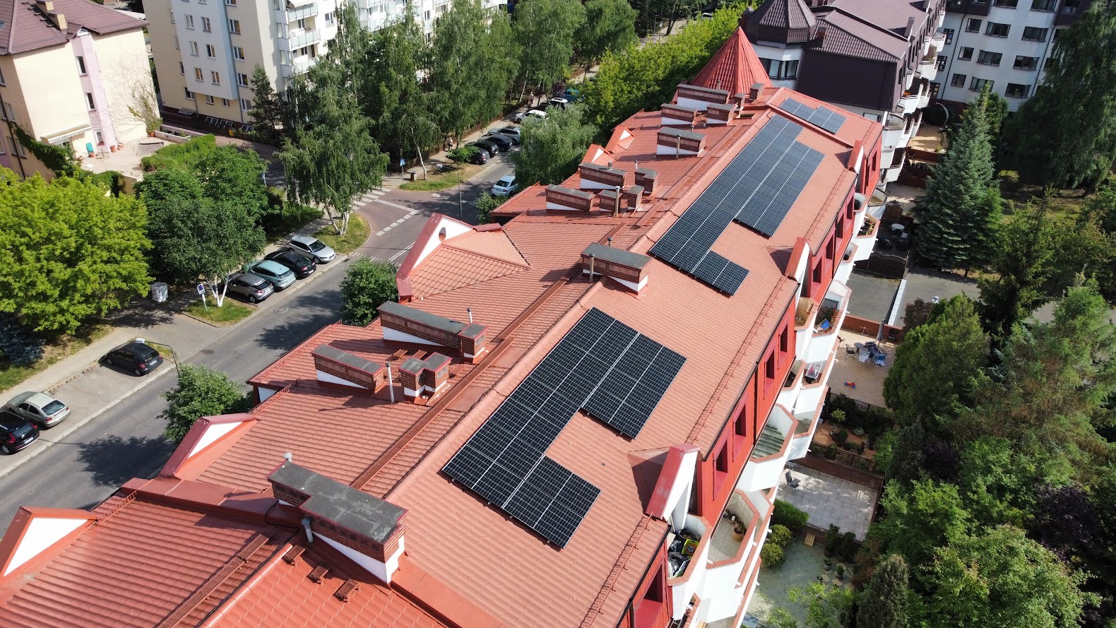 Widok z góry na budynek mieszkalny z czerwonym dachem, na którym zamontowano panele słoneczne w kilku sekcjach, otoczony zielenią i zabudową miejską.