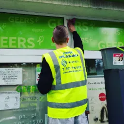 Pracownik firmy sprzątającej w odblaskowej kamizelce czyści okno lokalu z zielonym napisem 'GEERS', używając fioletowej ściereczki; obok stoi szare wiadro.