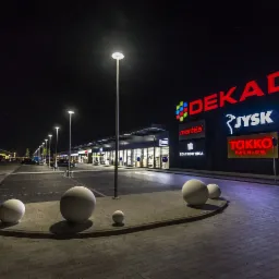 Nocny widok na oświetlony budynek centrum handlowego z logo różnych marek, kuliste dekoracje na pierwszym planie i rzędem lamp ulicznych wzdłuż parkingu.