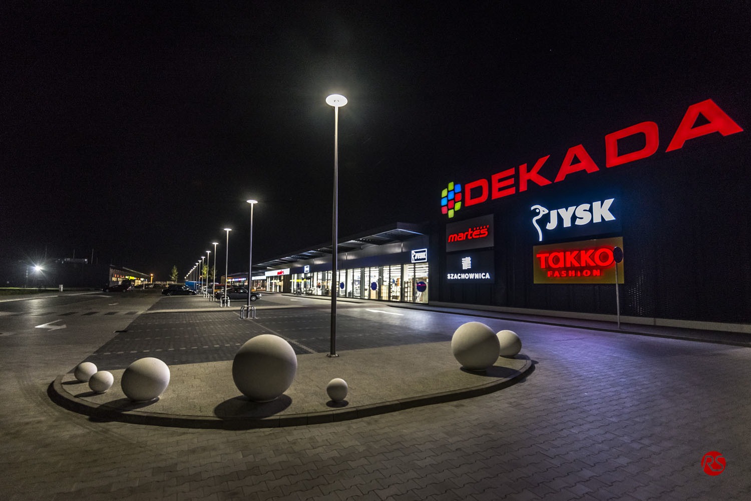 Nocny widok na oświetlony budynek centrum handlowego z logo różnych marek, kuliste dekoracje na pierwszym planie i rzędem lamp ulicznych wzdłuż parkingu.