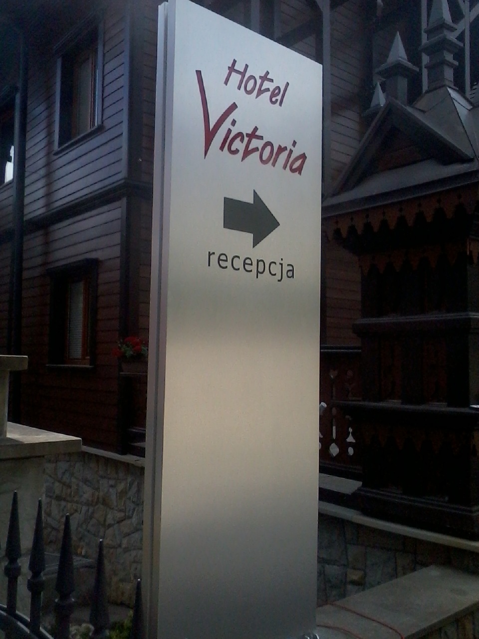 Pionowy szyld reklamowy Hotelu Victoria z napisem i strzałką wskazującą recepcję, umieszczony przed budynkiem w stylu zakopiańskim.
