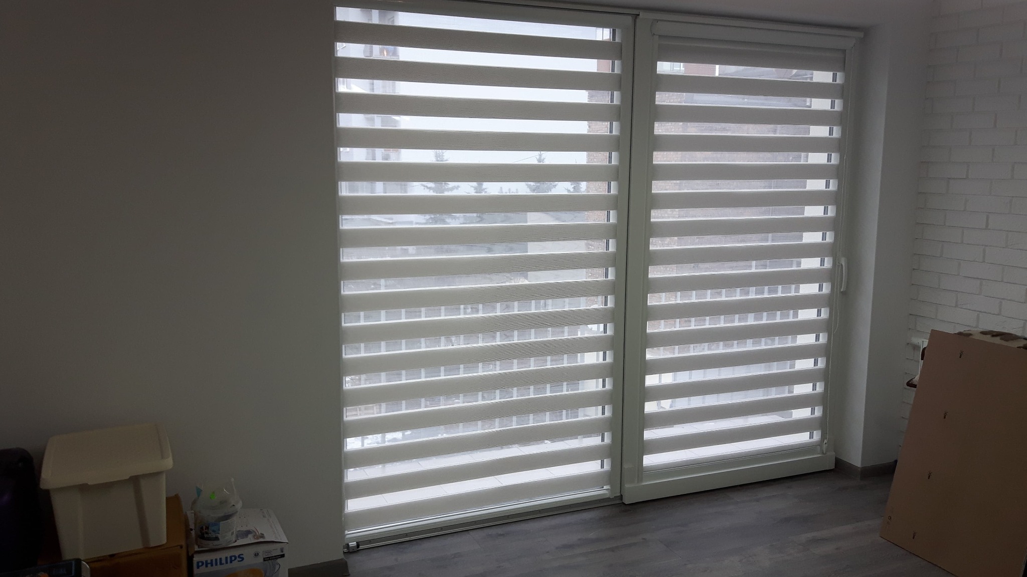 Dwa okna balkonowe z zamontowanymi, częściowo zasłoniętymi przesłonami okiennymi z poziomymi pasami, na jednym otynkowana ściana, na drugim ściana z białej cegły.