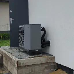 Szara pompa ciepła zamontowana na podwyższeniu z cegieł i kamiennej płyty, z widocznymi czarnymi rurami przy ścianie budynku i szarymi drzwiami wejściowymi w tle.