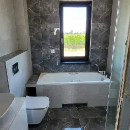 Nowoczesna łazienka z szarymi płytkami imitującymi marmur na ścianach i podłodze, białą wanną pod oknem z widokiem na zielone pole, umywalką na białej szafce i toaletą.