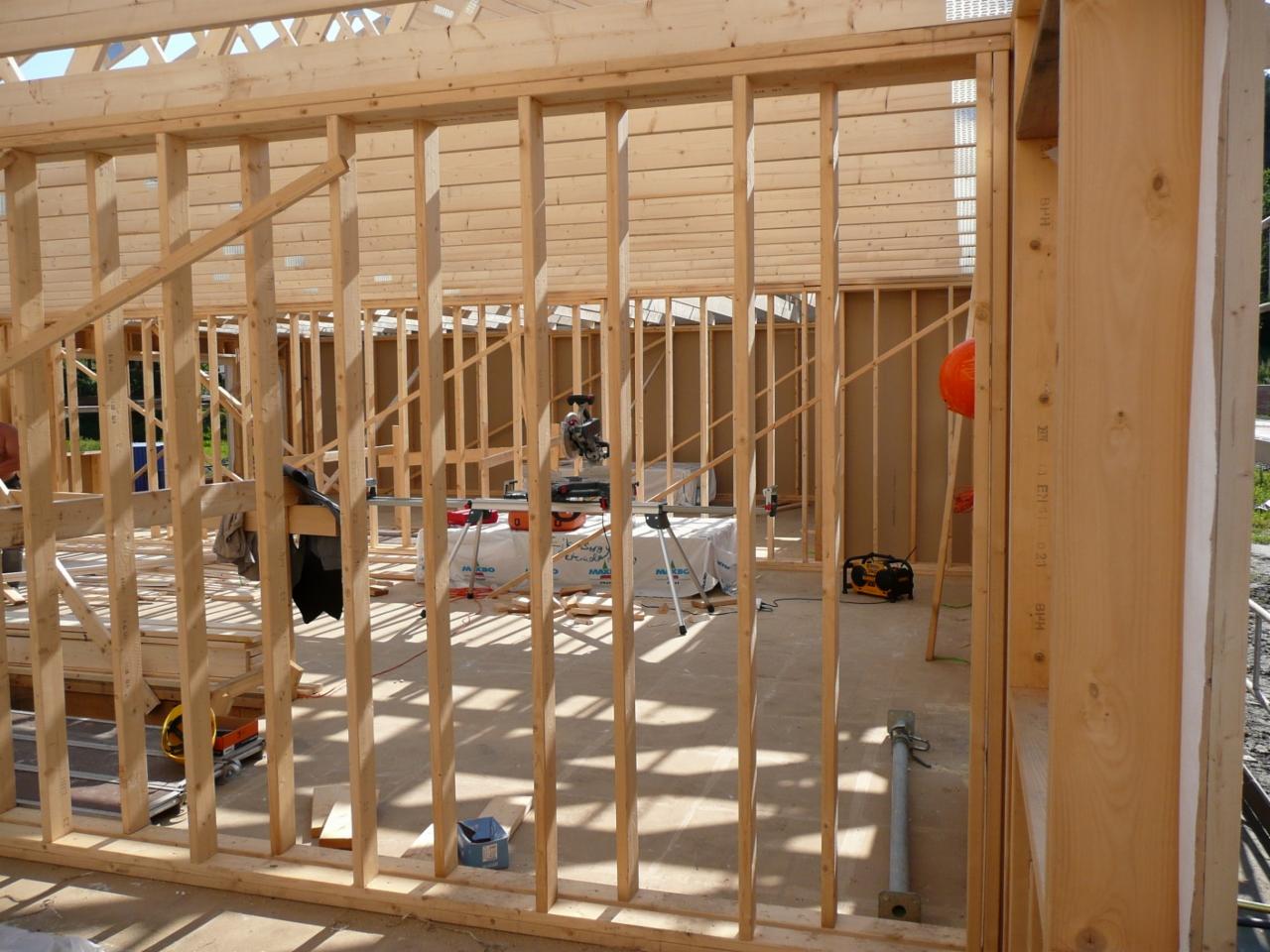 Surowy szkielet drewniany budynku w trakcie budowy, widoczne narzędzia i materiały budowlane w tle, pomarańczowy kask na ścianie.