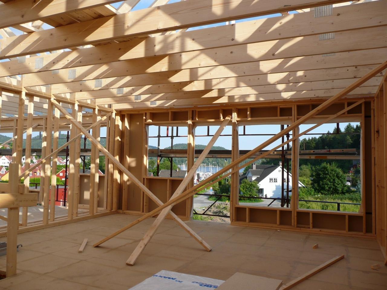 Wnętrze surowego stanu budynku z drewnianą konstrukcją szkieletową, widok przez okna na okoliczne domy i jezioro.