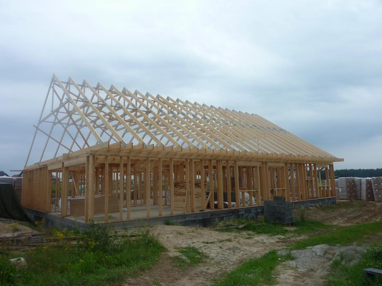 Konstrukcja drewniana domu w trakcie budowy, widoczne ściany szkieletowe i więźba dachowa na fundamencie, w tle pochmurne niebo.