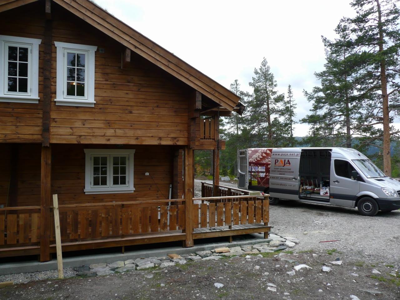 Dwukondygnacyjny, drewniany dom z bali z białymi ramami okiennymi i balustradą werandy; w tle las iglasty i furgonetka z logo firmy.