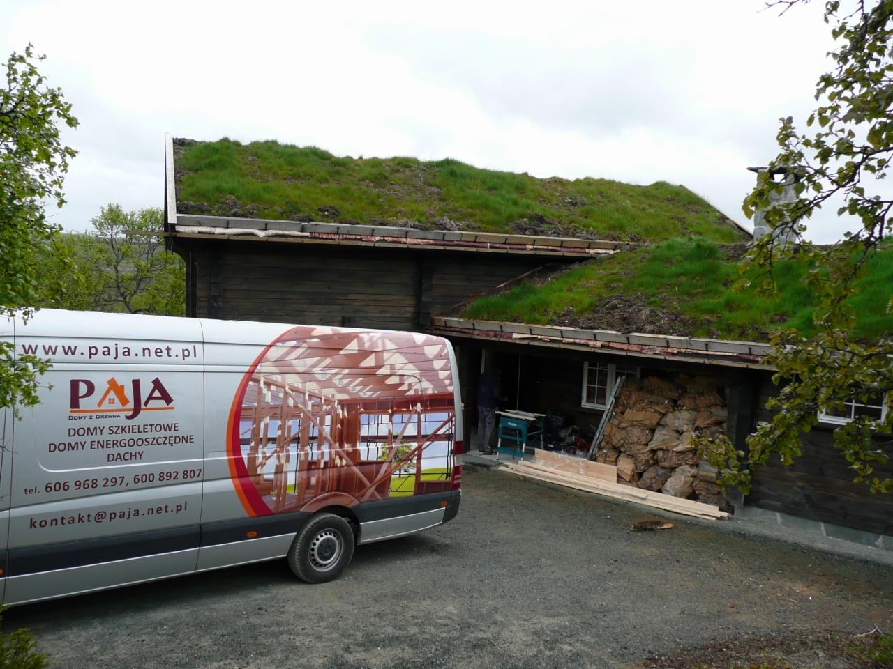Ciemny drewniany budynek z zielonym dachem porośniętym trawą, częściowo zasłonięty przez samochód dostawczy z logo firmy budowlanej i wizualizacją konstrukcji szkieletowej.
