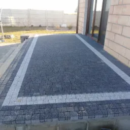 Zakończona realizacja tarasu z ciemnej kostki brukowej z jasnymi obramowaniami, widoczny zagęszczarka w tle, dom z beżowej cegły i betonowe ogrodzenie.