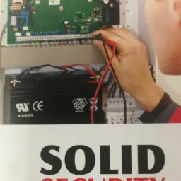 Instalator sprawdza napięcie w centrali alarmowej z podłączonym akumulatorem, widoczna płyta główna i okablowanie.