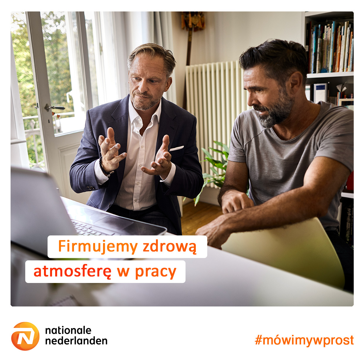 Dwóch mężczyzn w trakcie rozmowy online na laptopie w jasnym biurze, jeden w garniturze gestykuluje, drugi w koszulce słucha, w tle regał z książkami i roślina doniczkowa.