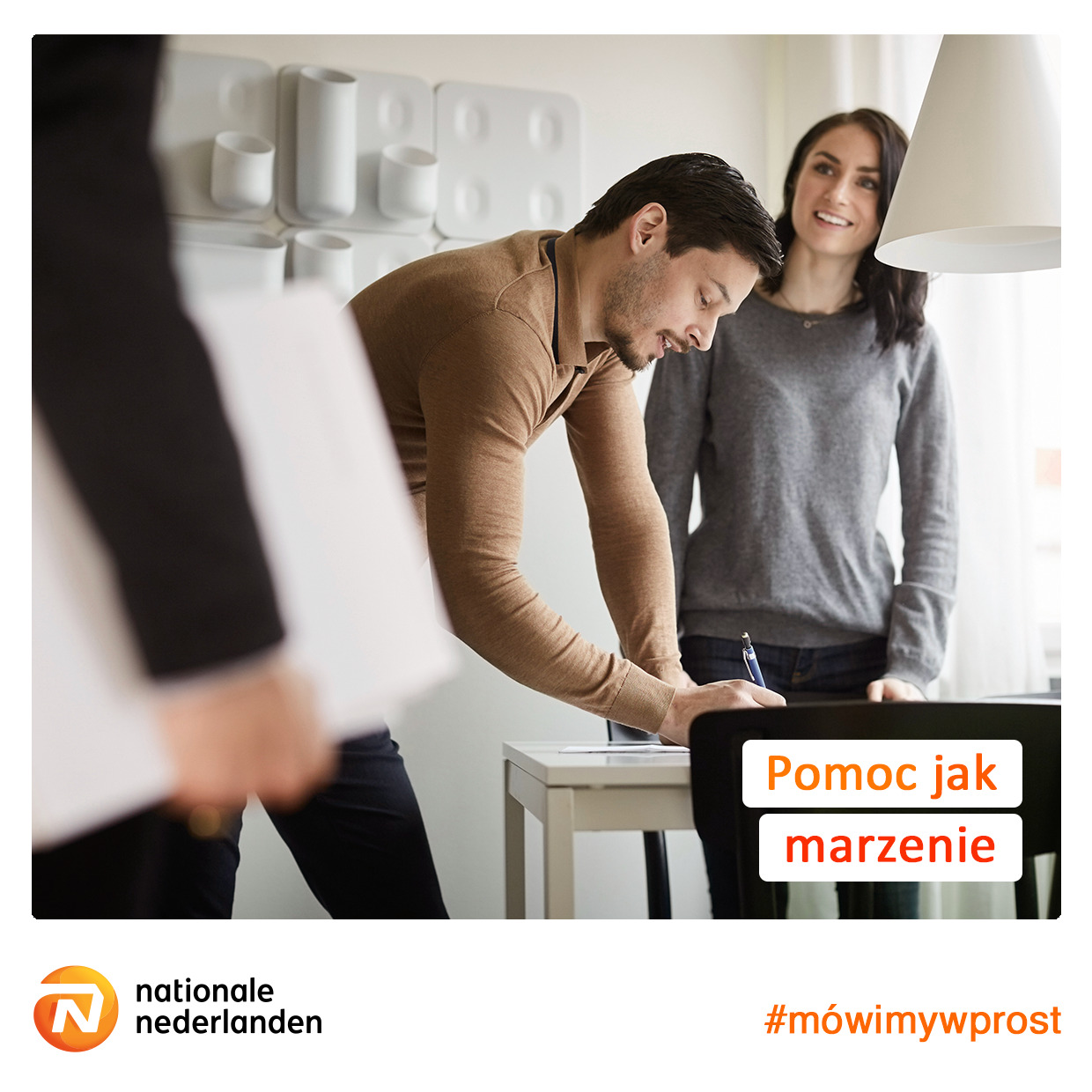 Mężczyzna podpisuje dokument przy biurku, kobieta stoi obok, w tle biała ściana z dekoracjami, widoczny fragment osoby trzymającej dokumenty.