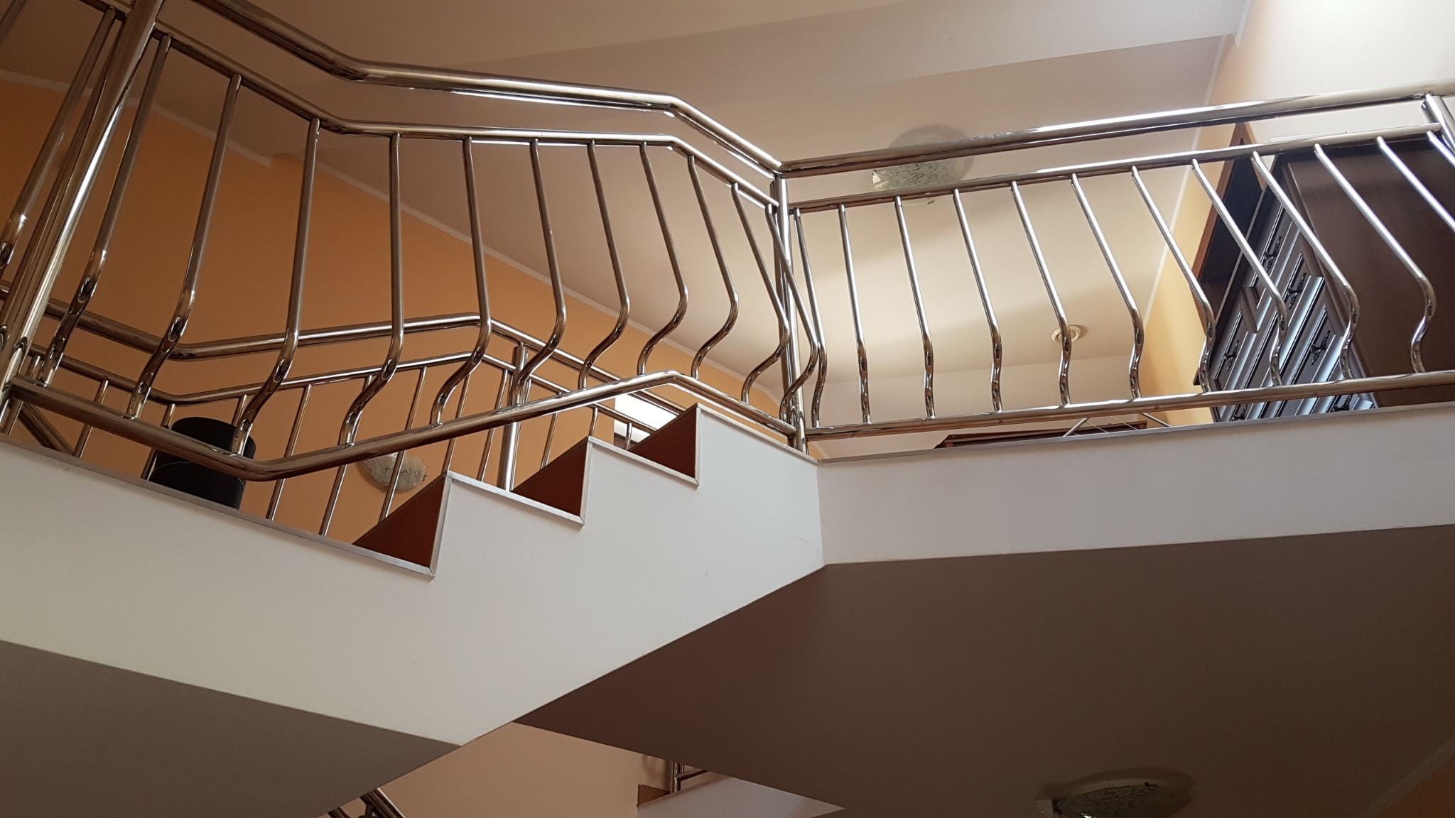 Wewnętrzne schody z białymi stopniami i balustradą ze stali nierdzewnej o falistych pionowych elementach, widok z dołu na górę.