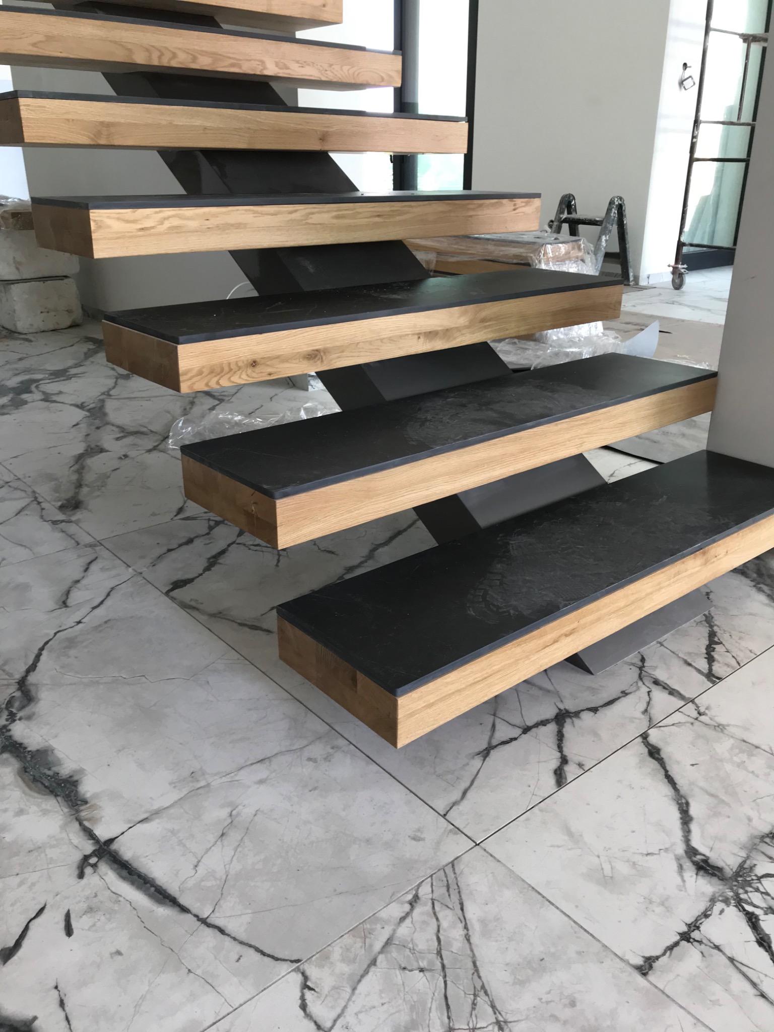 Nowoczesne schody z drewnianymi stopniami o czarnym wykończeniu, wsparte na metalowej konstrukcji, widok z dołu na tle marmurowej podłogi i drabiny w tle.