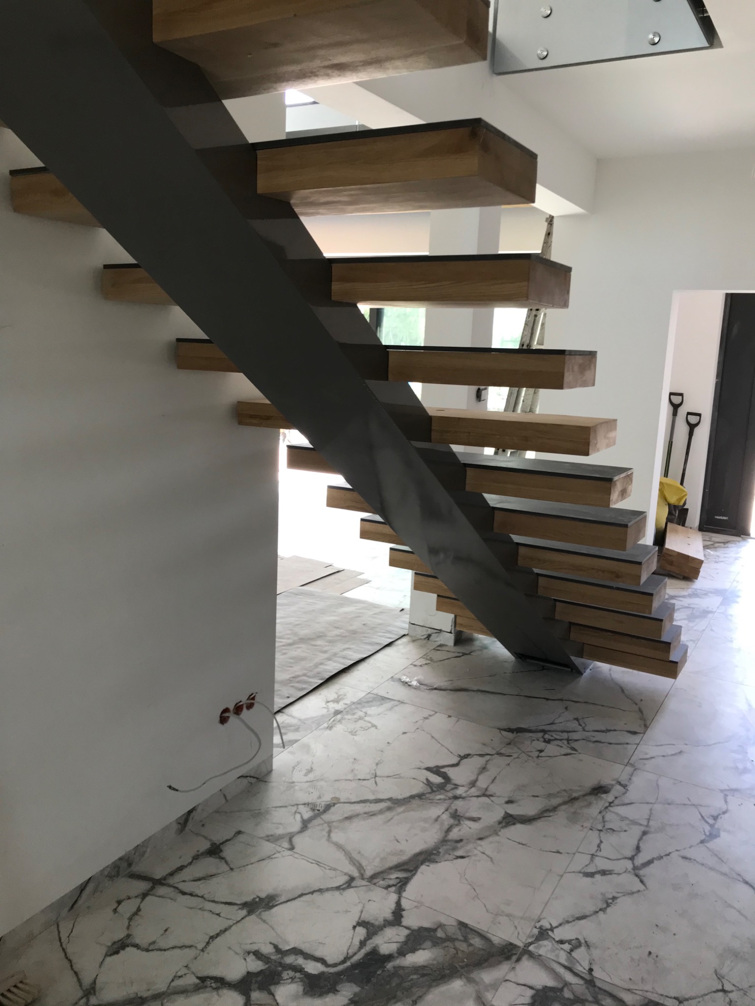 Nowoczesne schody wspornikowe z drewnianymi stopniami i stalową konstrukcją nośną, widoczne na tle marmurowej podłogi i białych ścian w trakcie wykończenia.