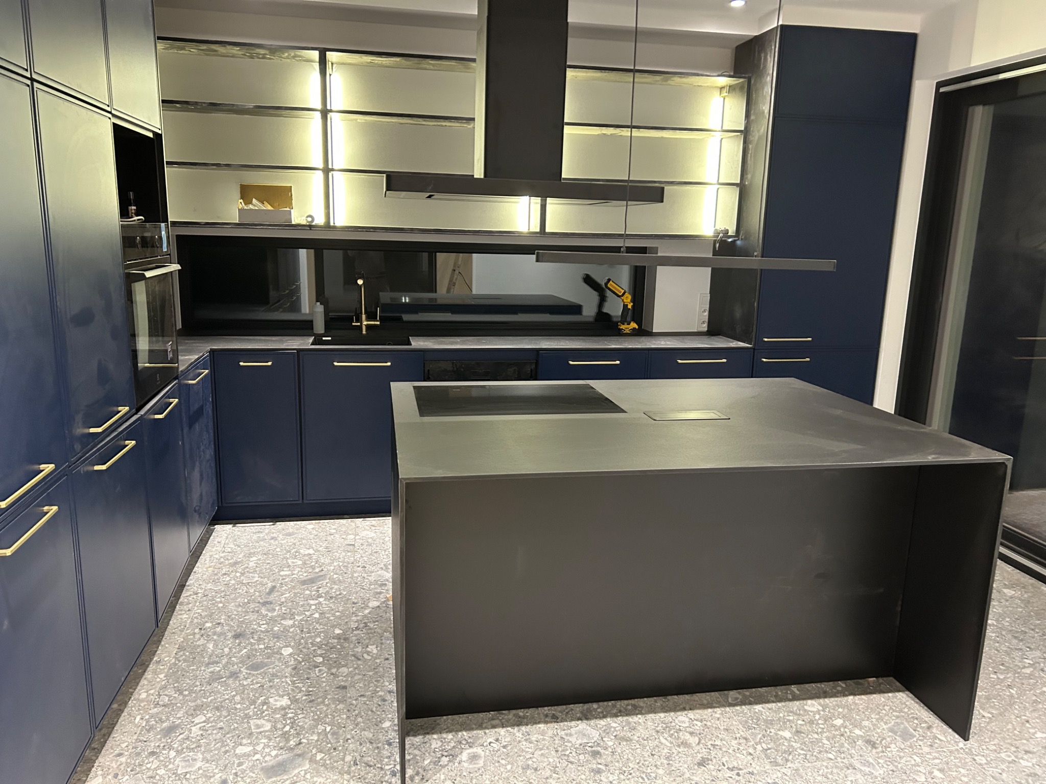 Nowoczesna kuchnia z ciemnymi, granatowymi szafkami z złotymi uchwytami, wyspą kuchenną w kolorze grafitowym, oświetleniem LED pod szafkami wiszącymi i szarymi płytkami na podłodze. W tle widoczny...